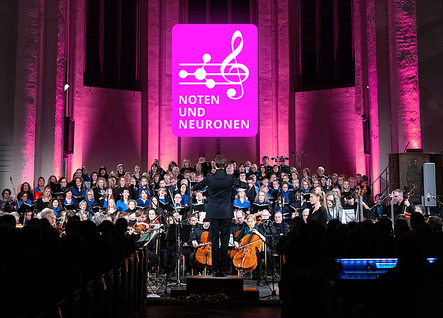 Ein Dirigent leitet ein großes Orchester und einen Chor in einem beleuchteten Konzertsaal, darüber ein Schild mit der Aufschrift „Noten und Neuronen“